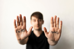 Eine unscharf abgebildete Person streckt die Handflächen Richtung Betrachter. Auf einer Hand steht das Wort Fake und auf der anderen News geschrieben.